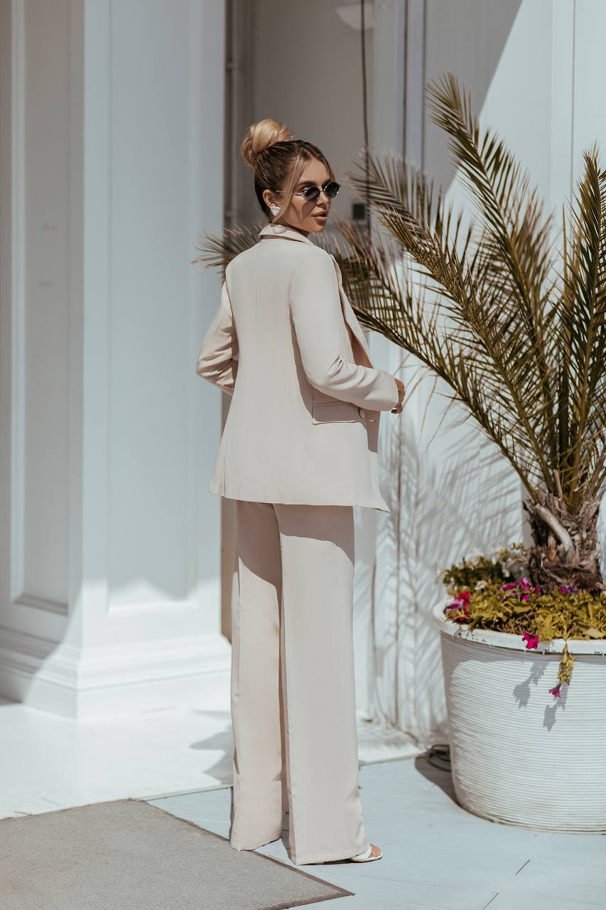 Woman in a beige suit standing next to a decorative plant and flower arrangement.