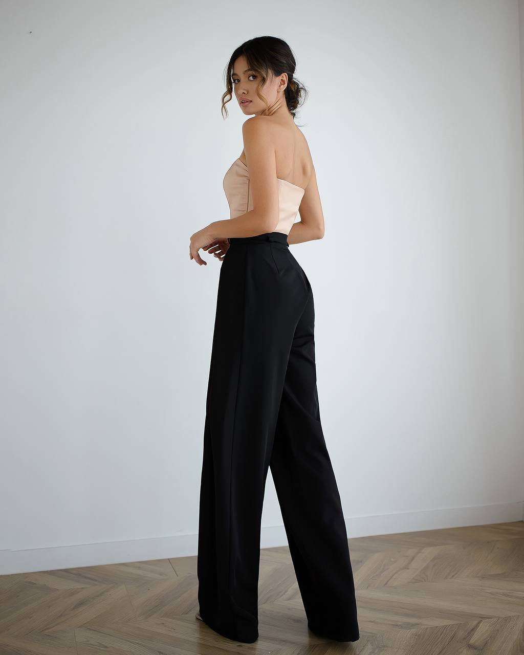 Woman wearing a strapless top and black wide-leg pants against a plain white wall.