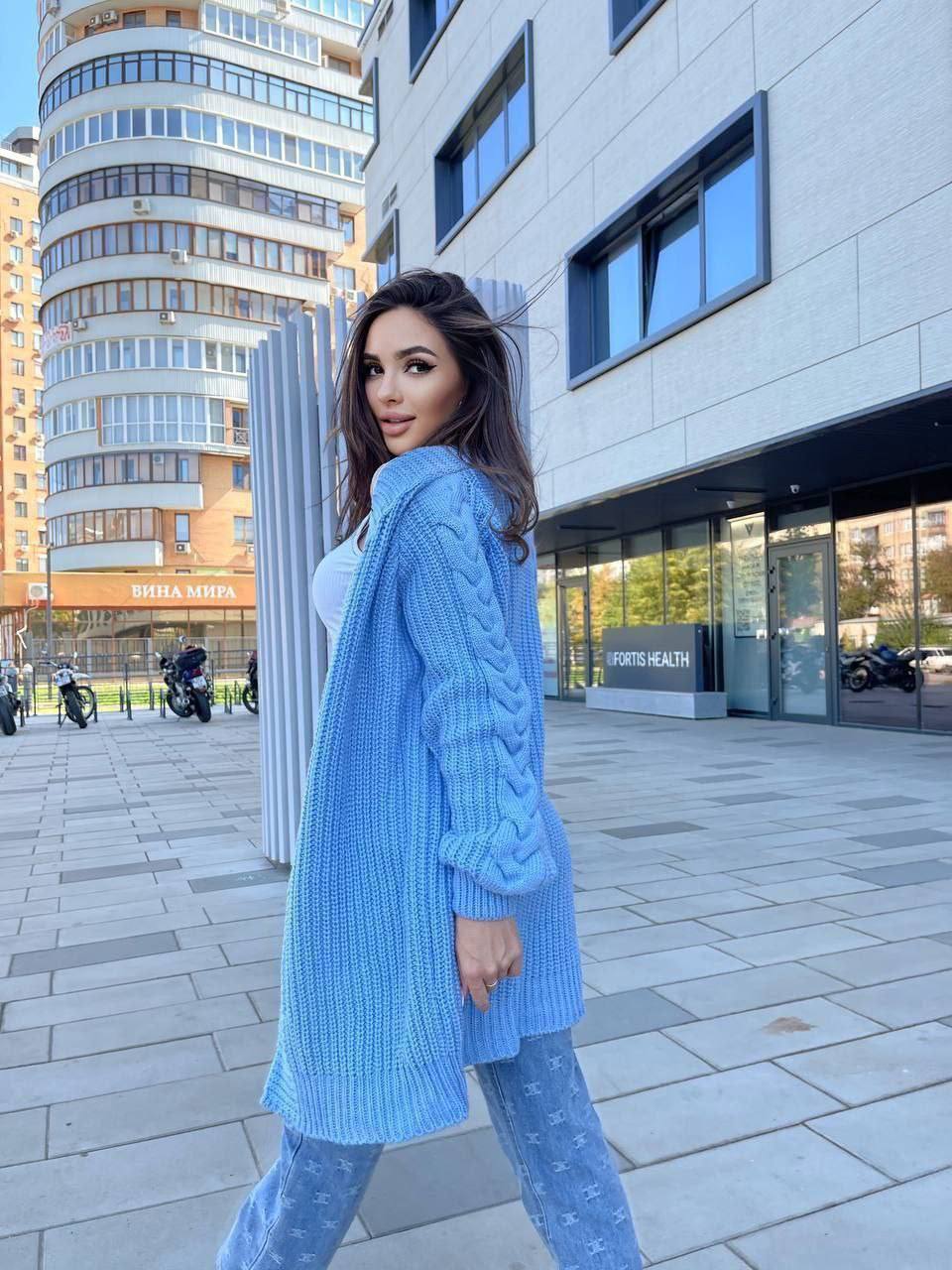 Woman wearing a blue knitted cardigan and pants standing on a city street.