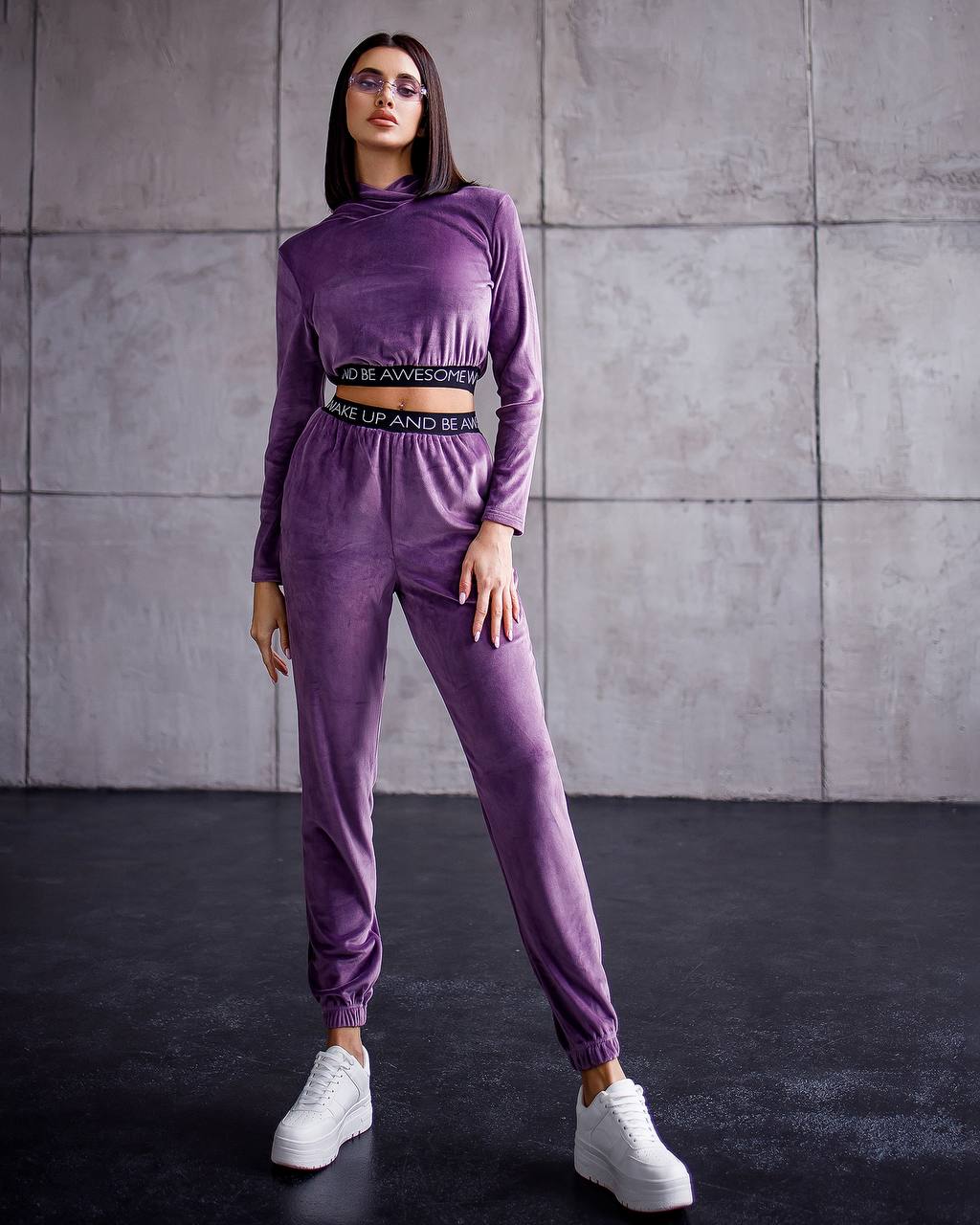Woman wearing a purple tracksuit with white sneakers against a gray tiled wall.