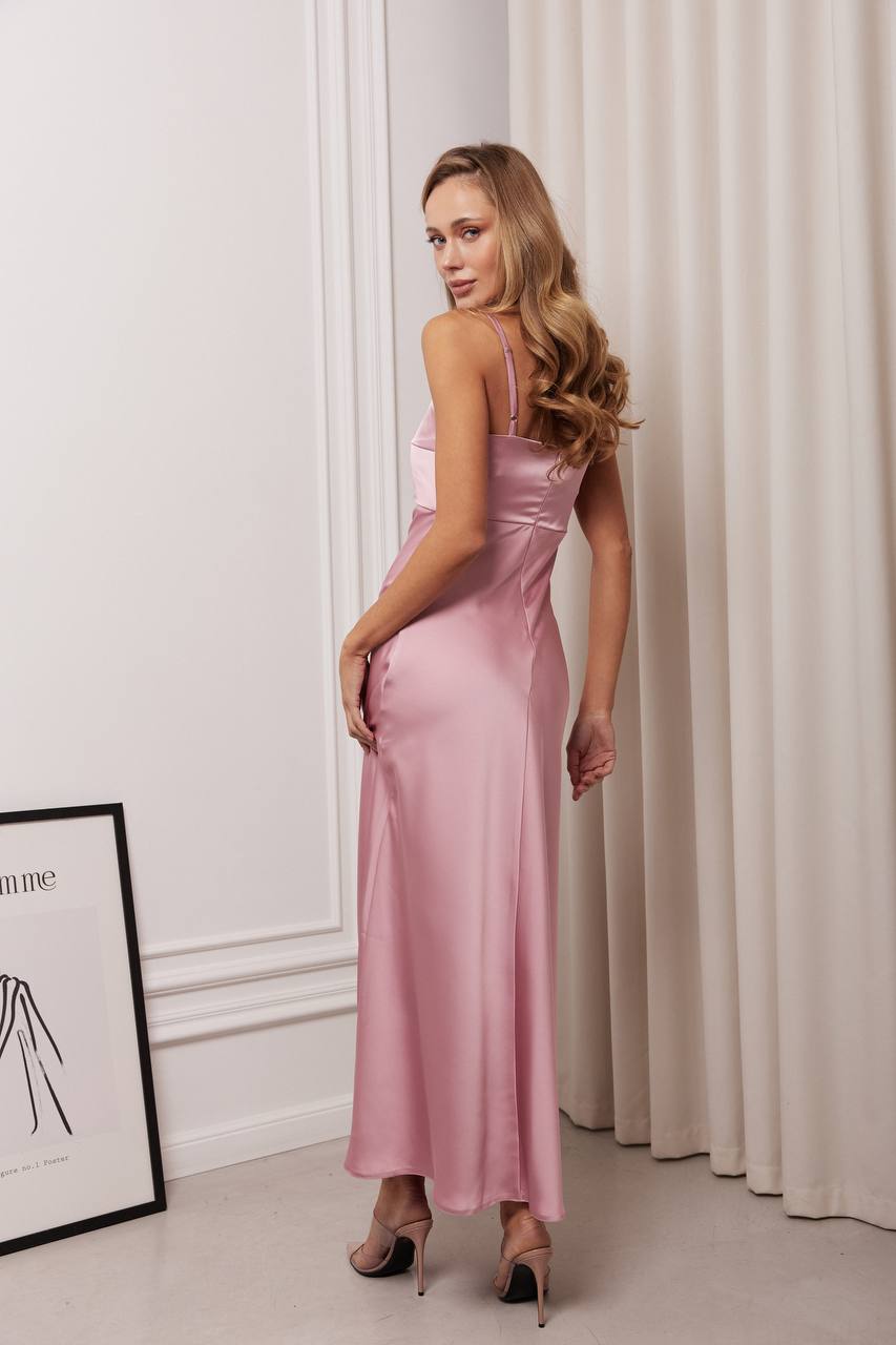 Woman wearing a pink satin dress standing against a white wall with curtains.