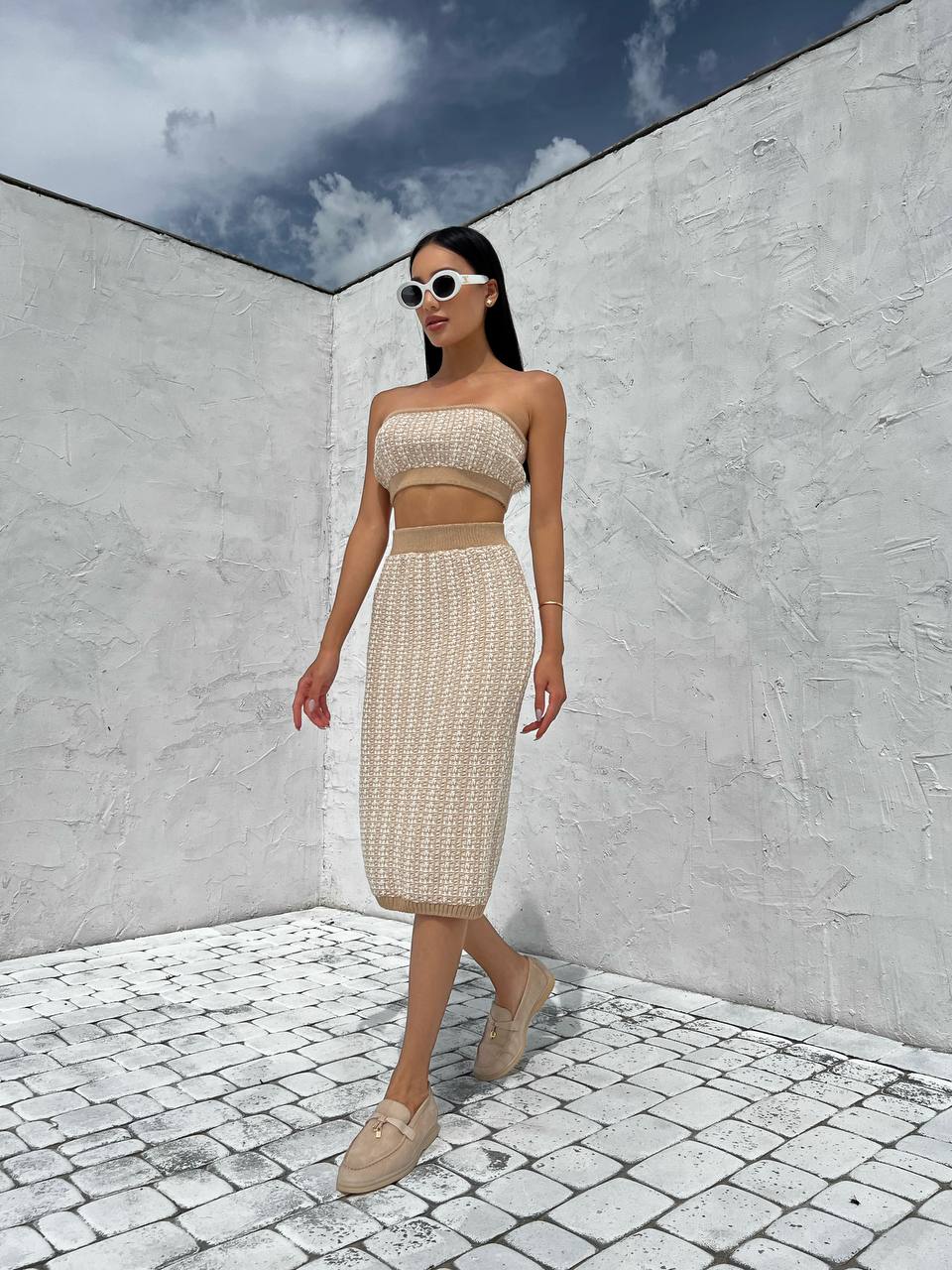 Woman in a beige outfit standing against a white wall with a blue sky.