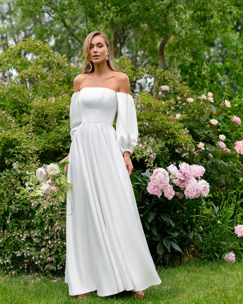 Woman in a white off-shoulder dress standing in a garden with flowers.