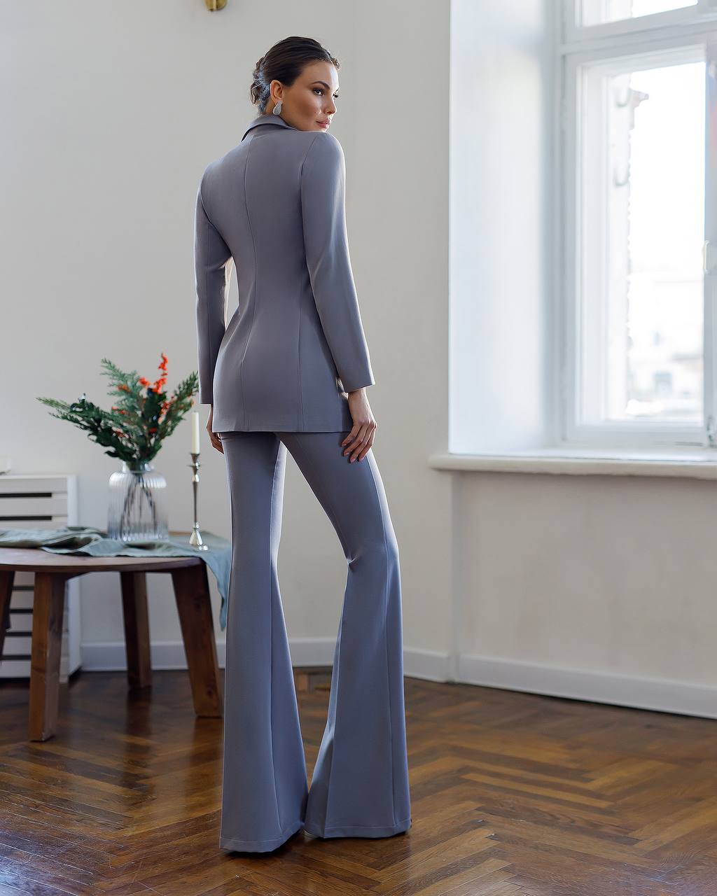 Woman in a gray suit standing in a room with a window and table.