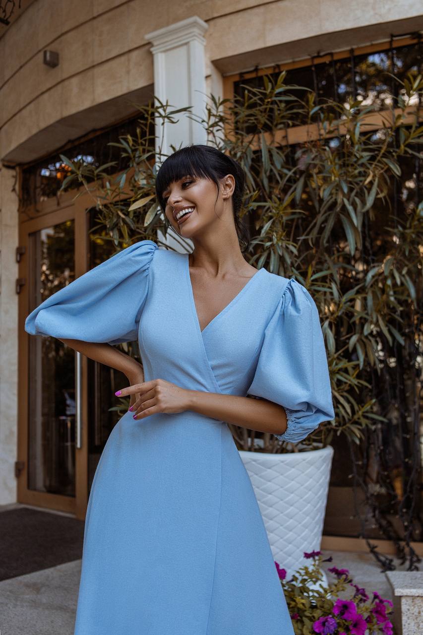 Woman in a light blue dress standing outdoors with plants and a building in the background