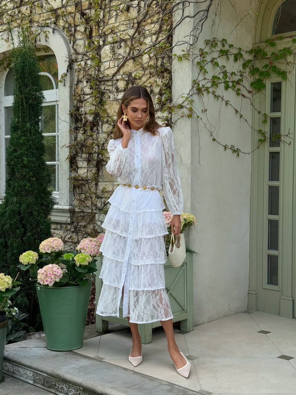 Woman in a white lace dress standing outdoors with flowers and a building in the background
