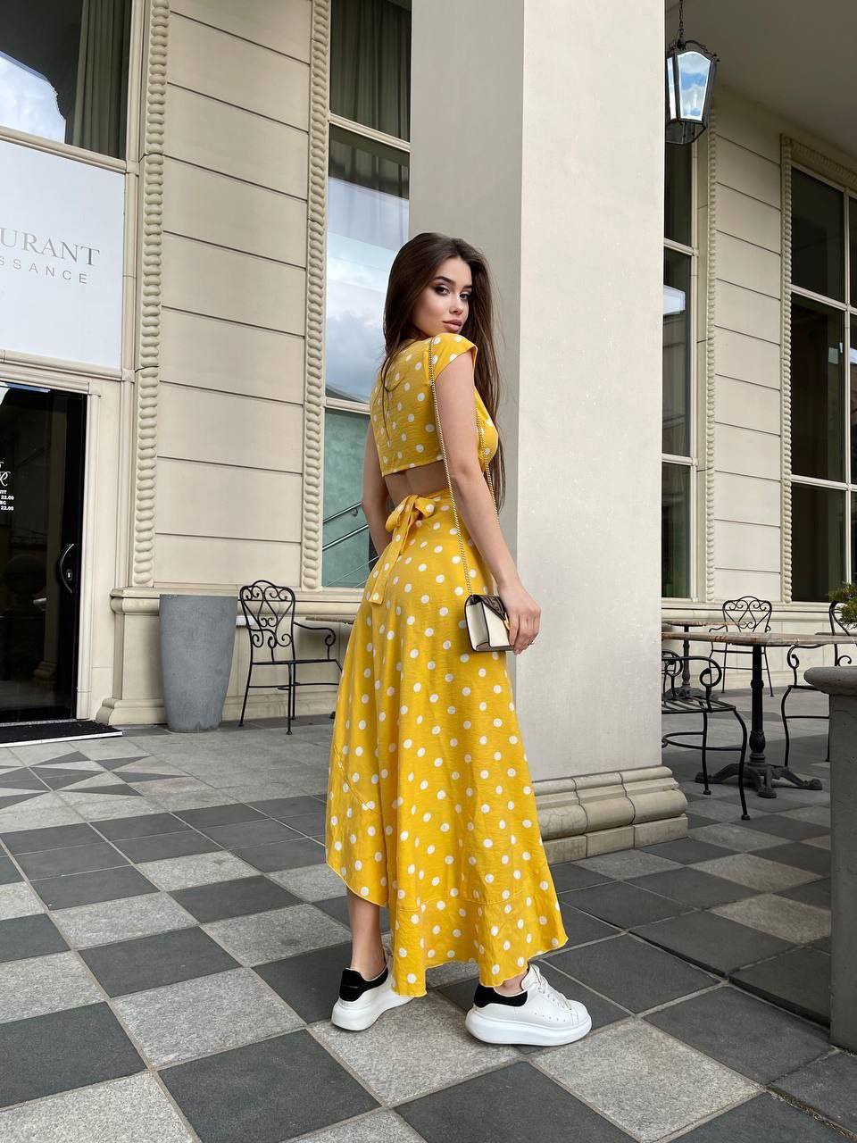 Woman in a yellow polka dot dress standing on a checkered pavement with a building in the background.