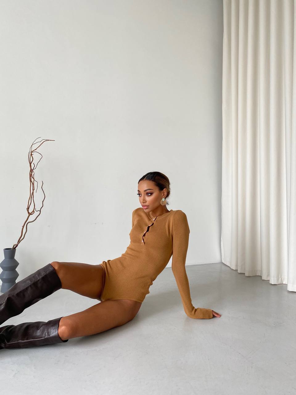 Woman in a mustard yellow dress and brown knee-high boots sitting on a light gray floor.