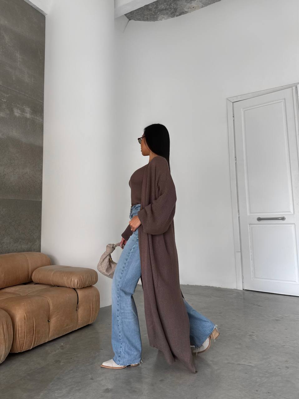 Person wearing a long brown coat and blue jeans standing in a minimalistic room with a brown sofa and white walls.