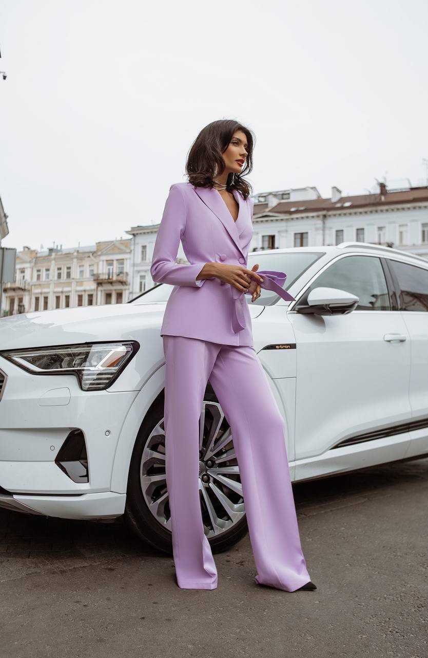 Woman in a lavender suit standing next to a white car with buildings in the background