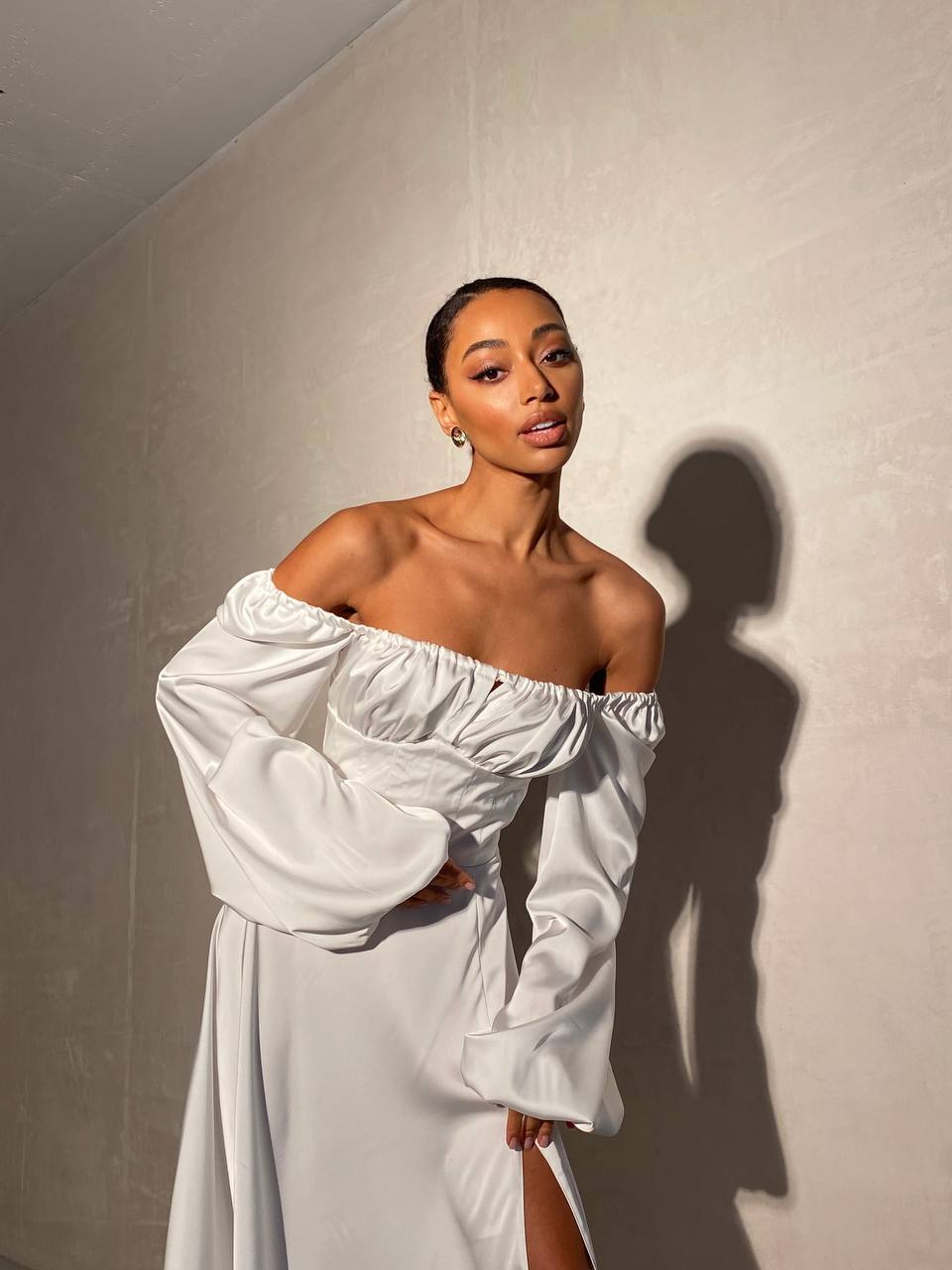 Woman wearing a white off-shoulder dress against a plain wall.