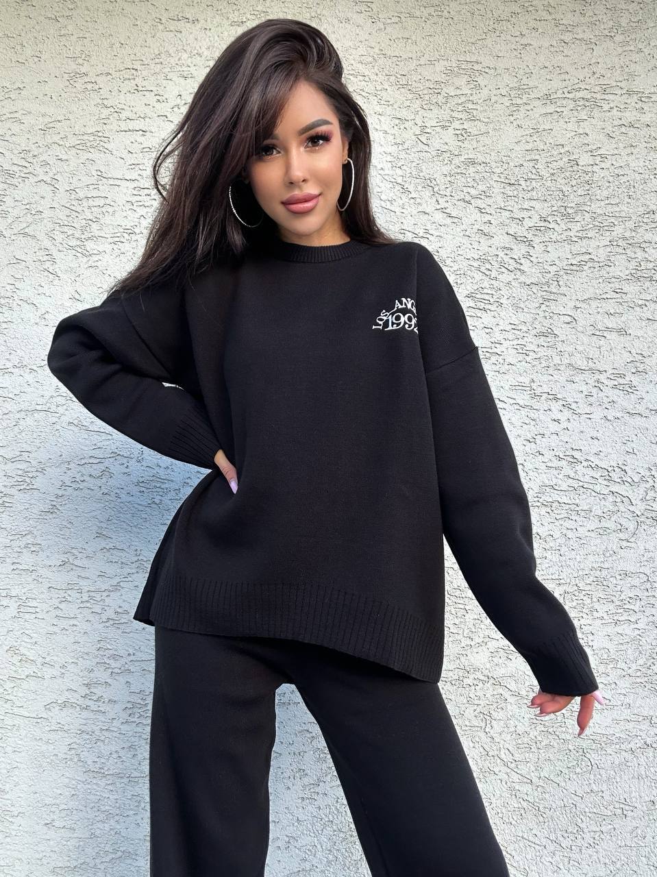Woman wearing a black sweater with a logo against a textured wall.