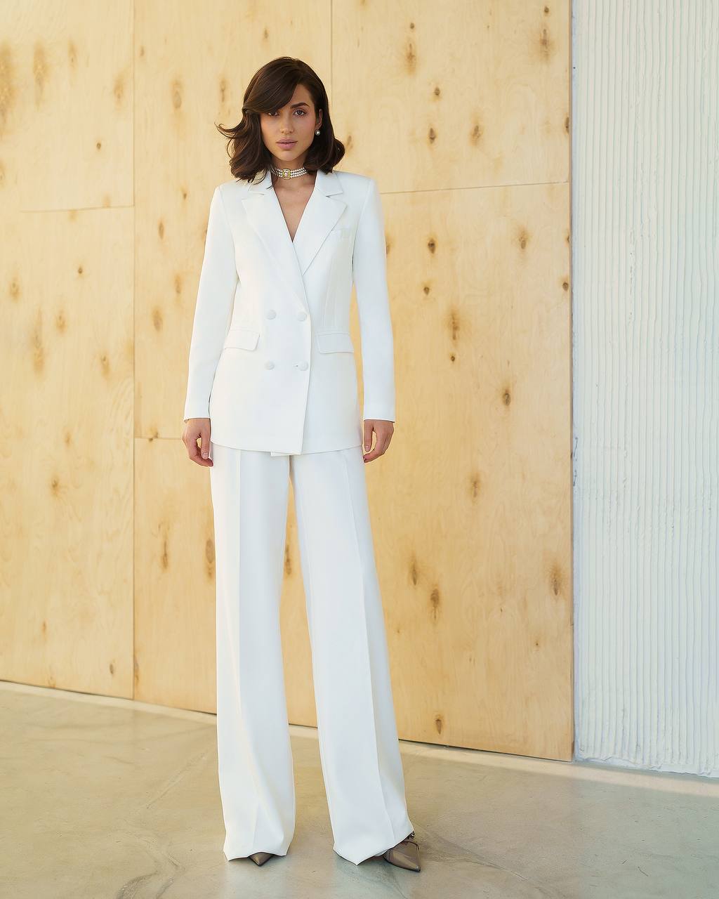 Woman wearing a white suit standing against a wooden wall.