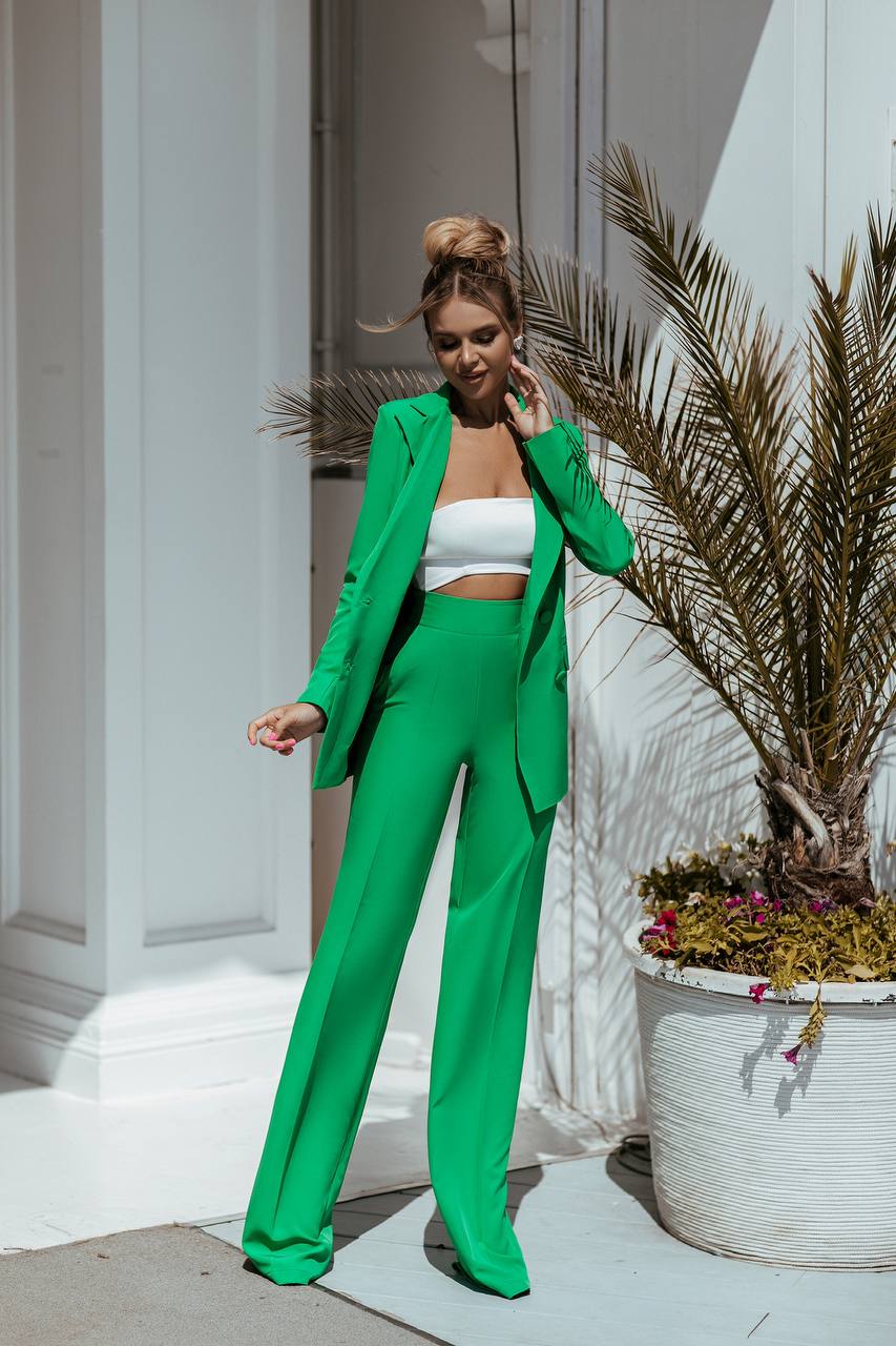 Woman in a green suit standing outdoors with plants and a building in the background