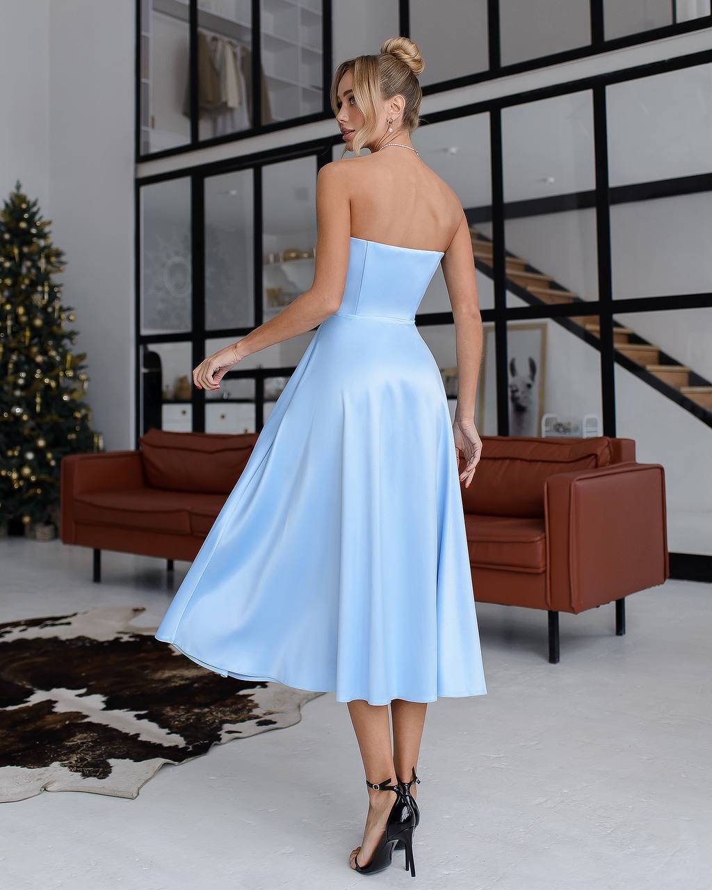 Woman in a light blue strapless dress standing in a modern room with brown sofas and a Christmas tree.