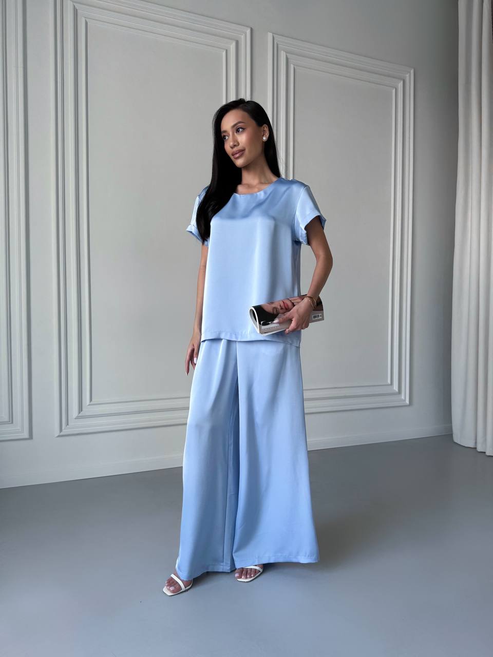 Woman wearing a light blue outfit standing against a white paneled wall.