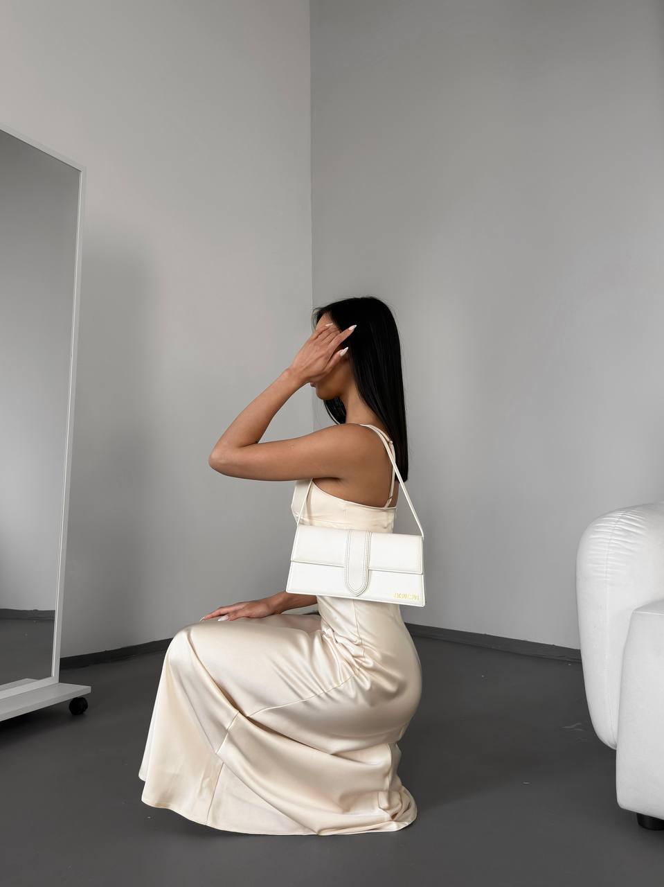 Woman in a white dress sitting on the floor with a white handbag, in a minimalistic room.