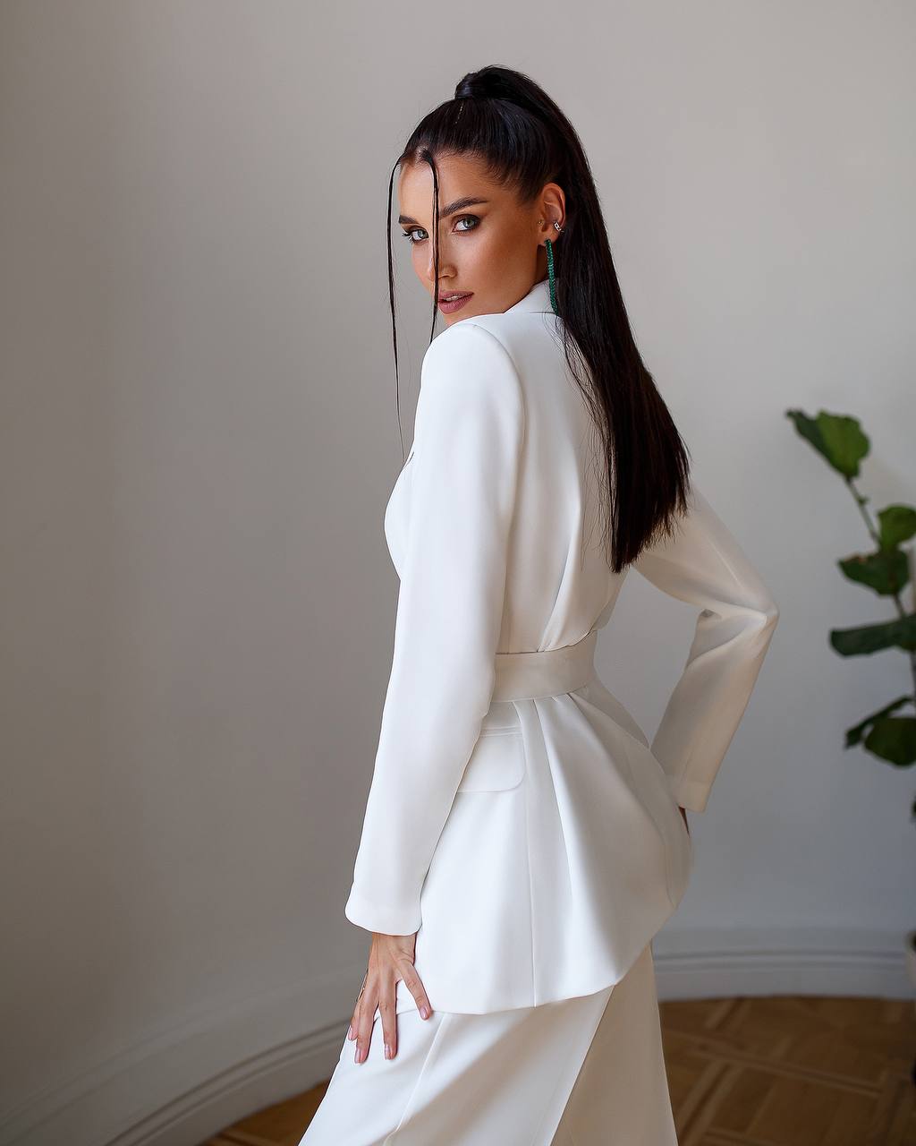 Woman wearing a white outfit standing against a plain wall with a plant in the corner.