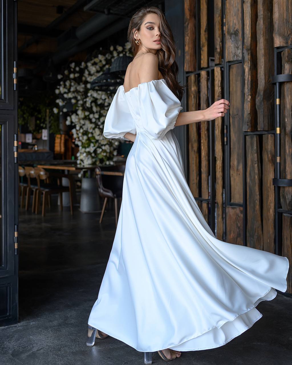 Woman in a white off-shoulder dress standing in a dimly lit room with wooden walls.