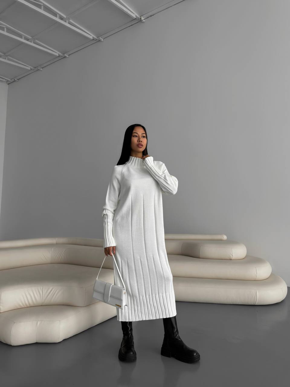 Woman in a white dress standing in a minimalistic room with gray walls and white furniture.