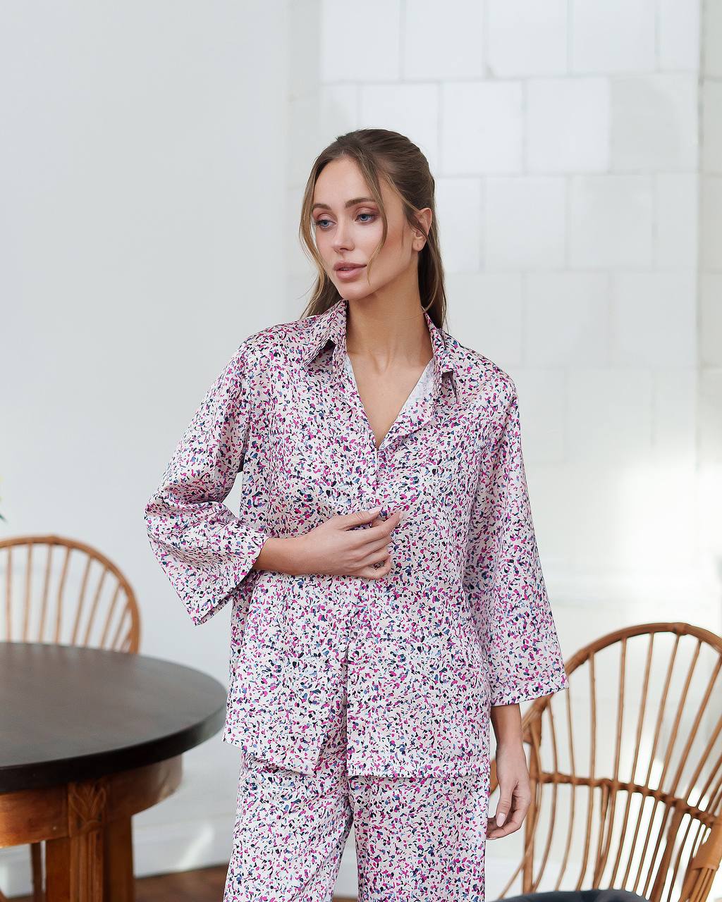 Woman wearing a pink floral pajama set in a room with wooden chairs and a table.