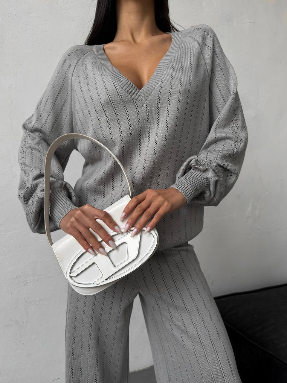 Person wearing a gray ribbed sweater and pants holding a white handbag against a gray background