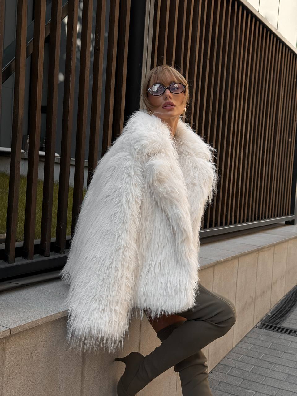 Person wearing a white fur coat and sunglasses against an urban background