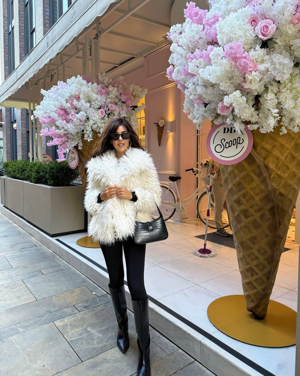 Woman in a white fur coat standing in front of a store with large floral displays and an ice cream cone sculpture.