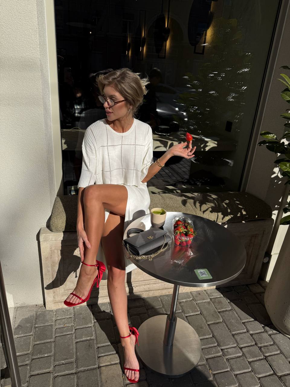 Woman sitting at a table outdoors, holding a phone, with a drink and a plant in the background.