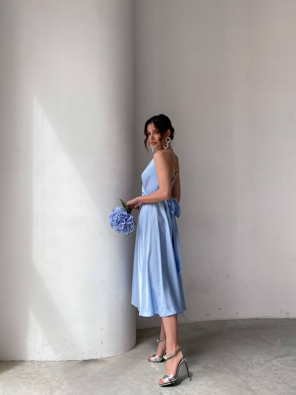 Woman in a light blue dress holding flowers against a plain wall.
