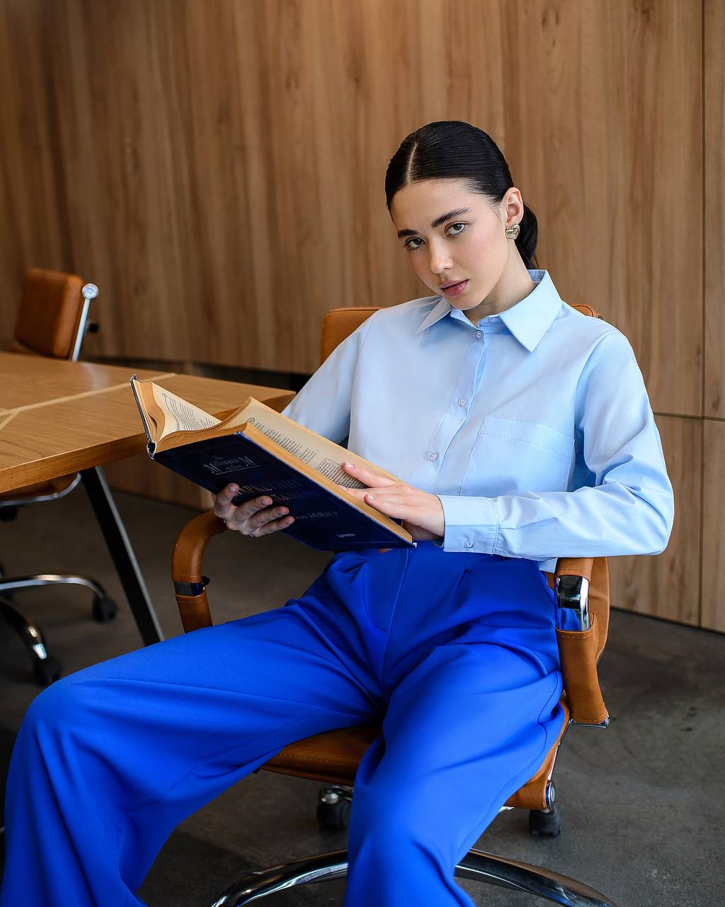Woman in a light blue shirt and blue pants sitting on a chair, holding an open book.