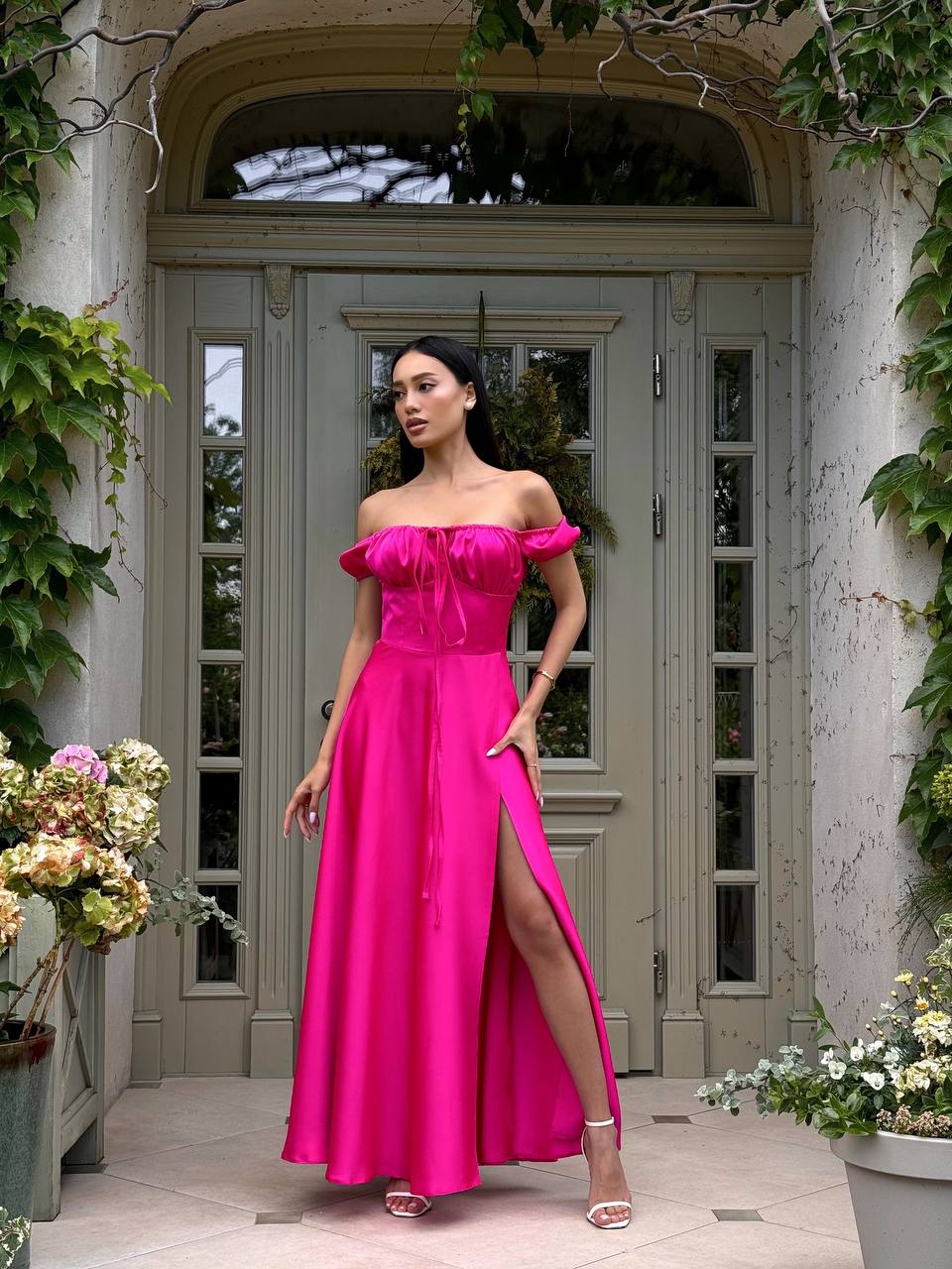 Woman in a bright pink off-shoulder gown standing in front of a gray door with greenery.