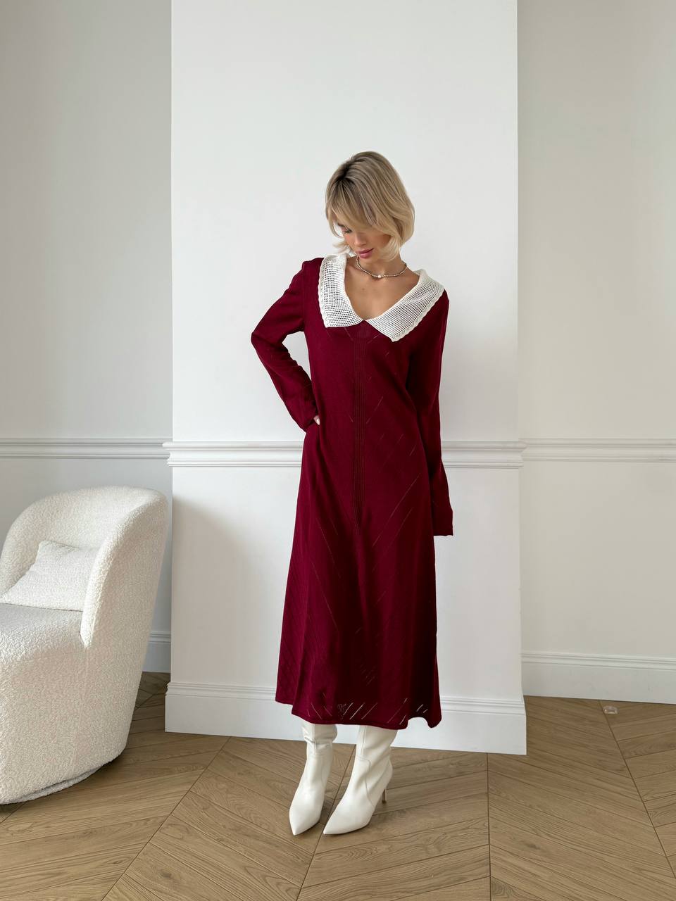 Woman wearing a burgundy dress with a white collar in a room with a white wall and wooden floor.