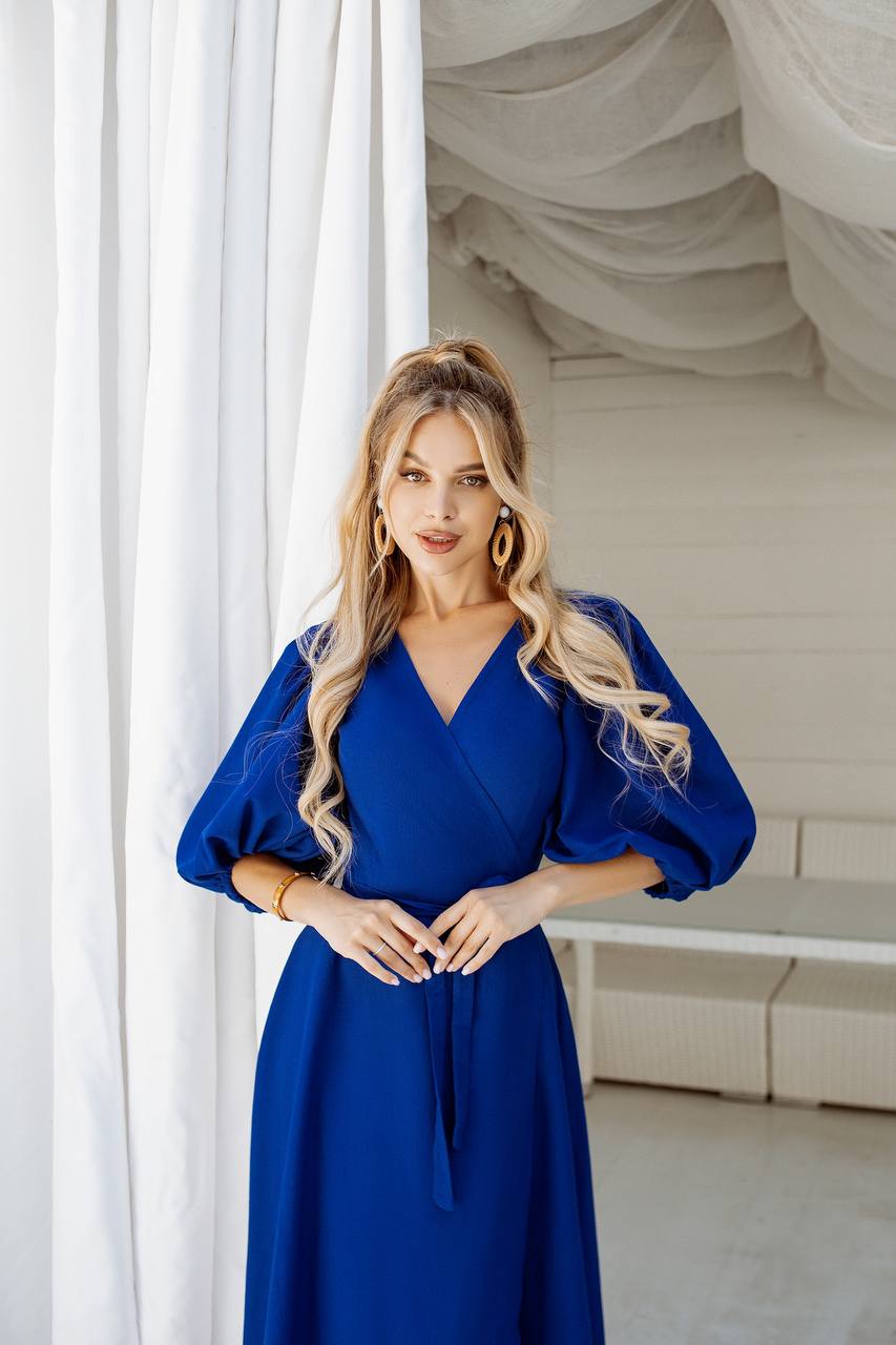 Woman in a blue dress standing in a room with white curtains.