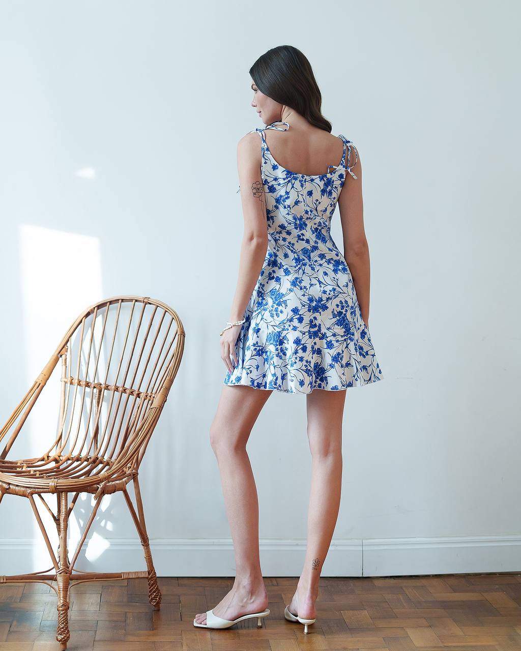 Woman wearing a blue floral dress standing next to a wicker chair indoors.