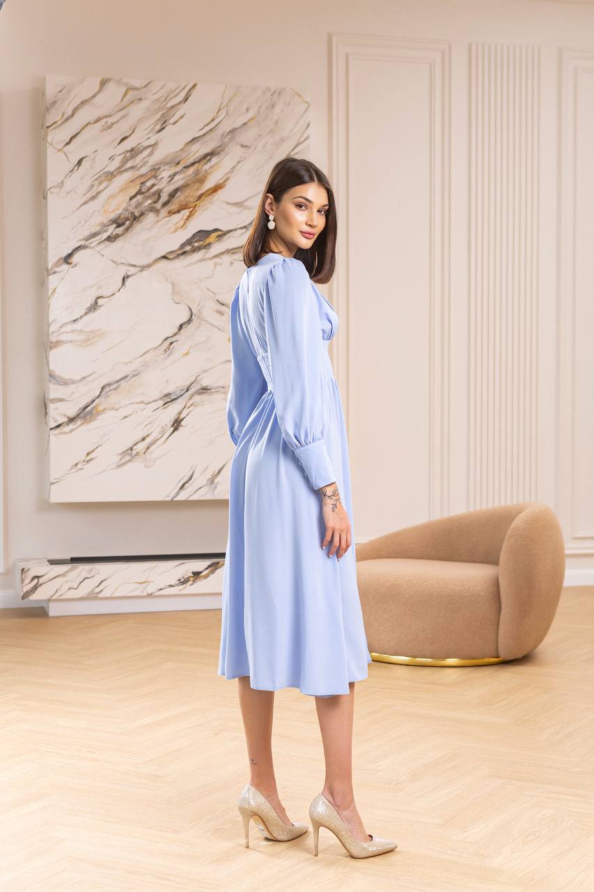 Woman in a light blue dress standing in a stylish room with a marble wall and beige armchair.