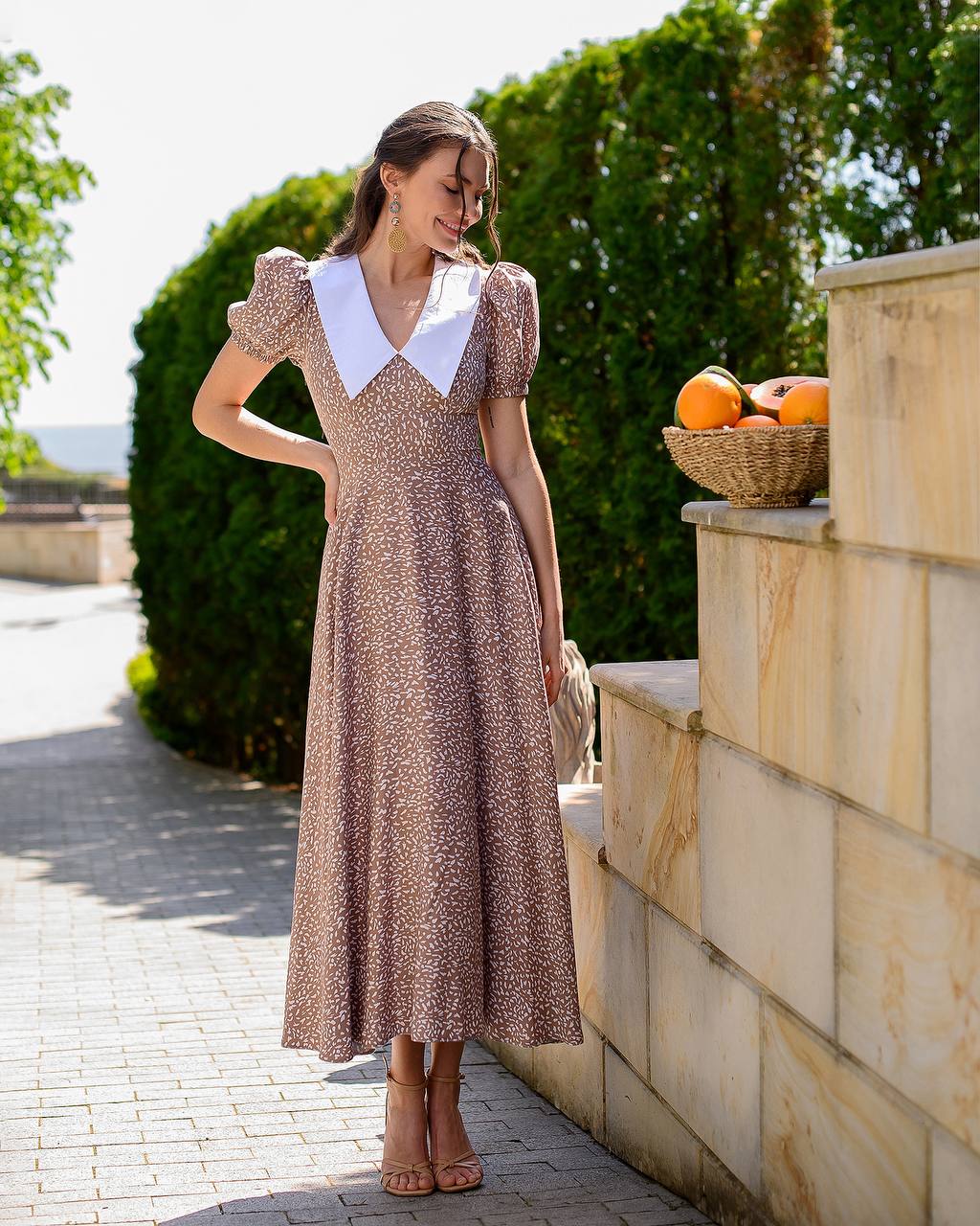 Woman in a vintage-style dress standing outdoors near a stone wall with a basket of oranges.