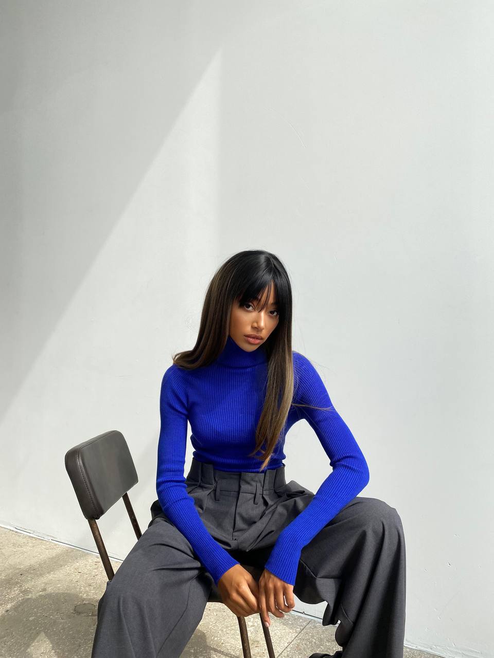 Person wearing a blue turtleneck and gray pants sitting on a chair against a white wall.