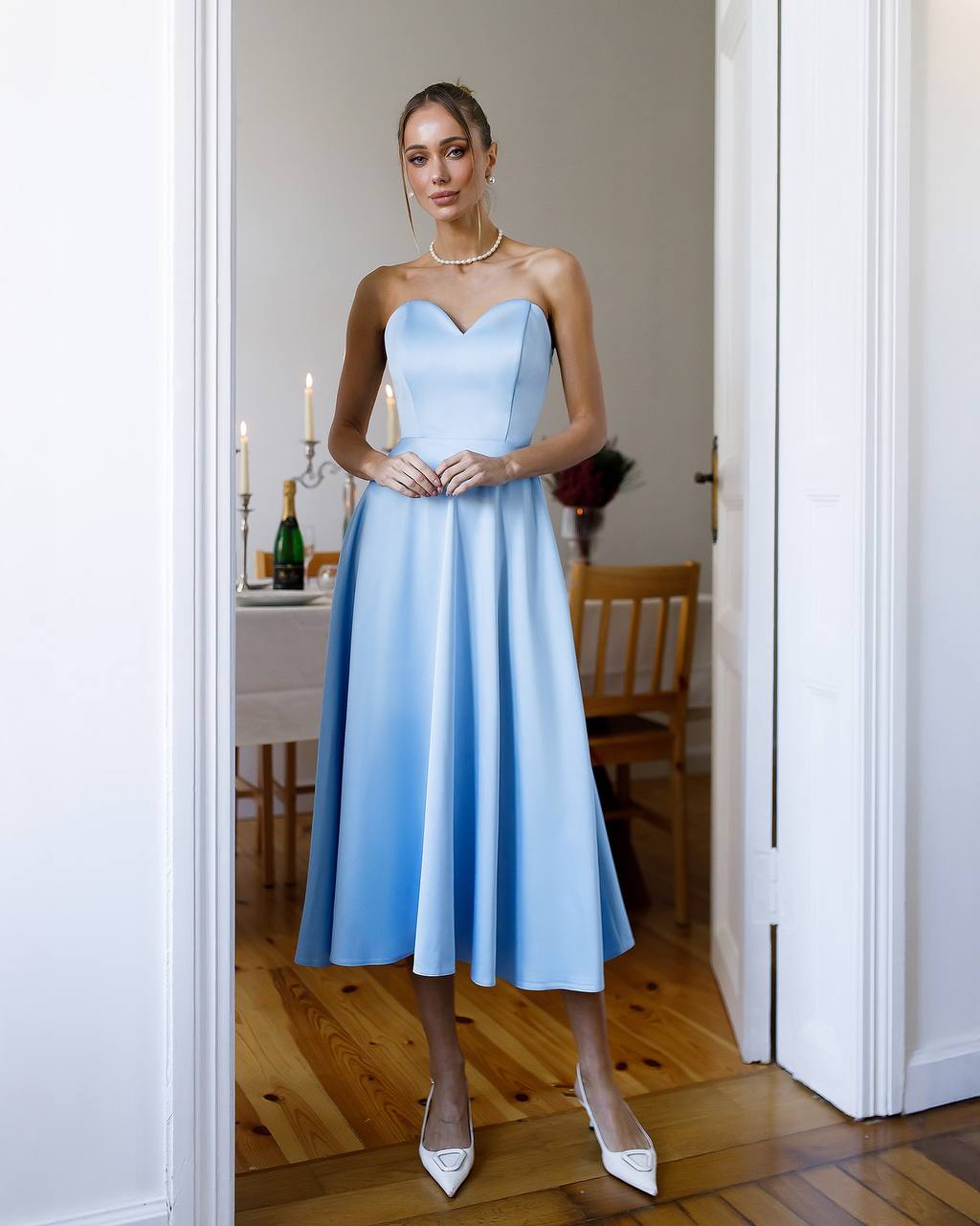 Woman in a light blue strapless dress standing in a room with wooden flooring and white walls.