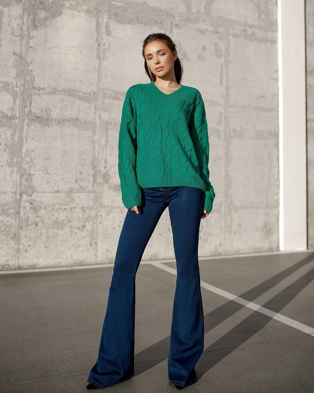 Woman wearing a green sweater and blue flared pants standing against a concrete wall.