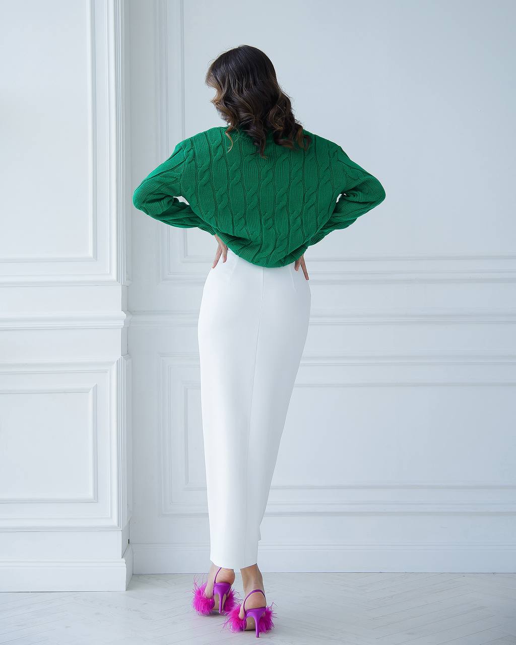 Woman wearing a green sweater and white pants standing against a white wall.