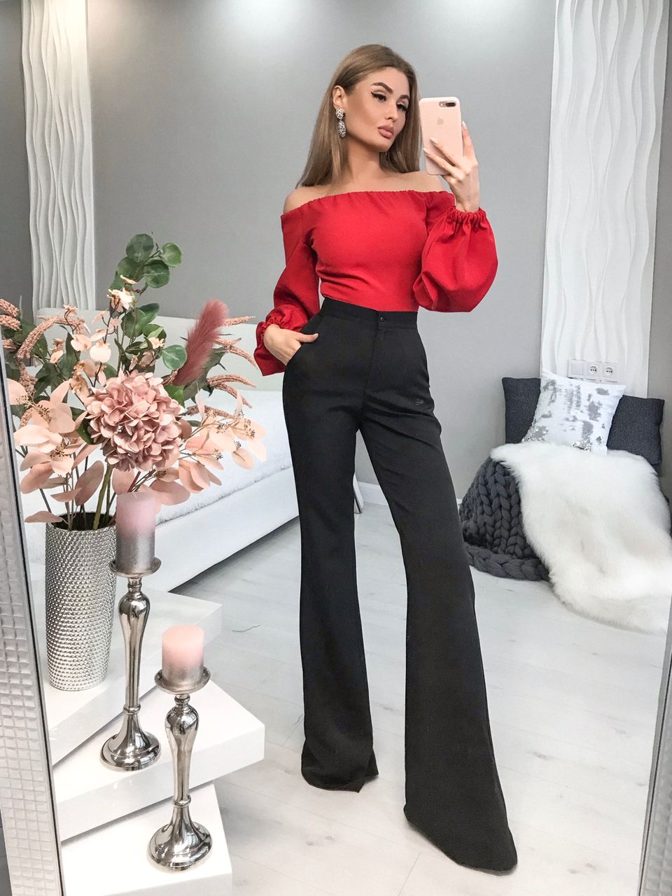 Woman taking a mirror selfie wearing a red top and black pants in a living room.