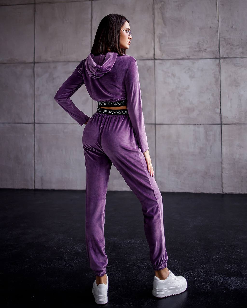 Woman wearing a purple tracksuit with a belt against a gray tiled wall.