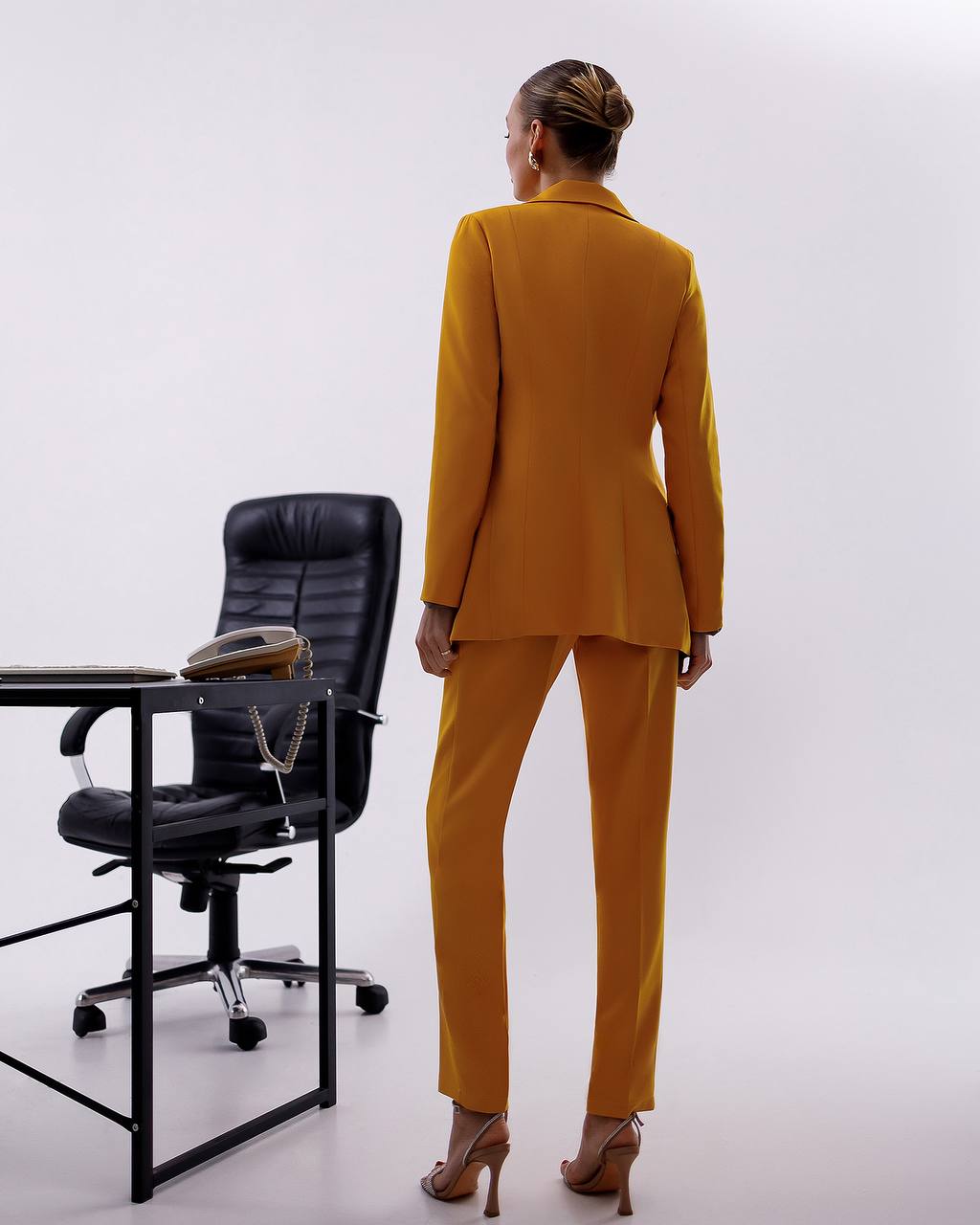 Woman in a mustard yellow suit standing in front of a black office chair on a white background