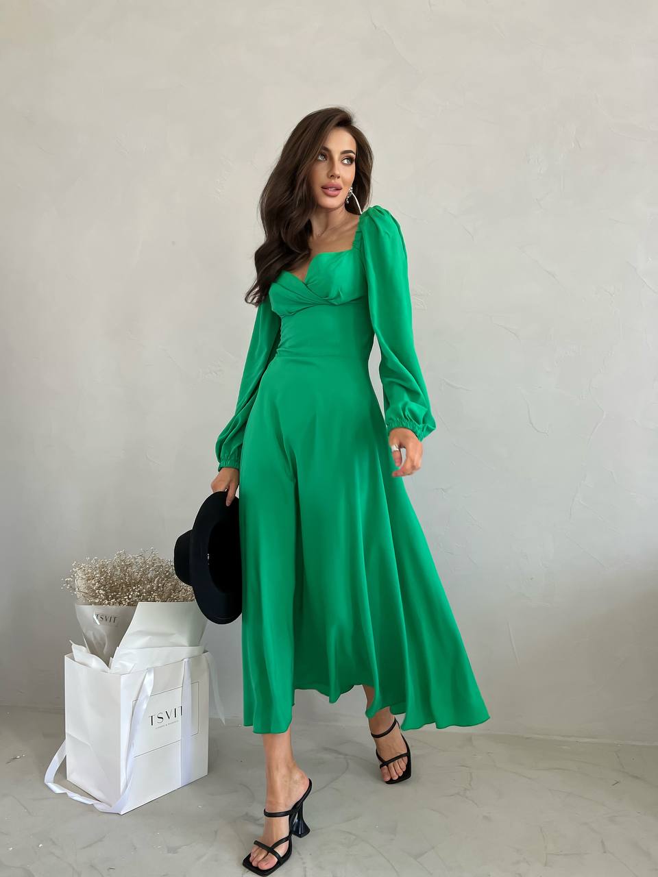 Woman in a green dress holding a black handbag against a plain background