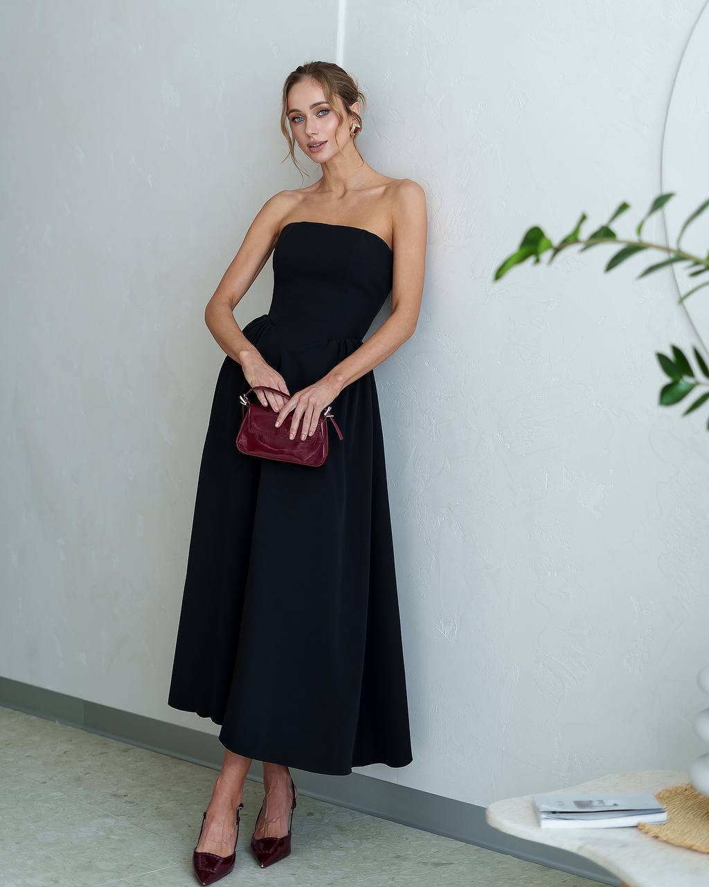 Woman in a black strapless dress holding a red clutch against a white wall.