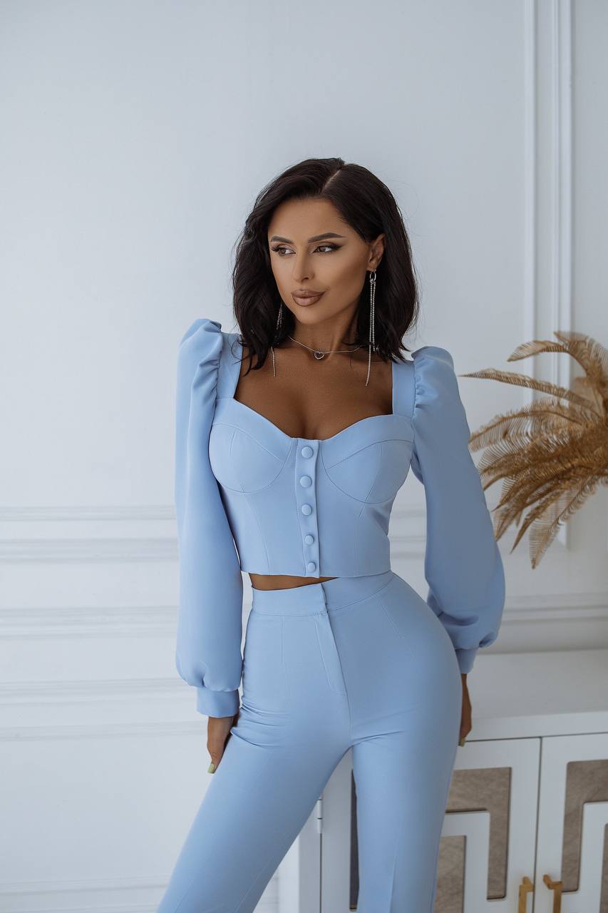 Woman wearing a light blue outfit standing against a white wall with decorative elements.