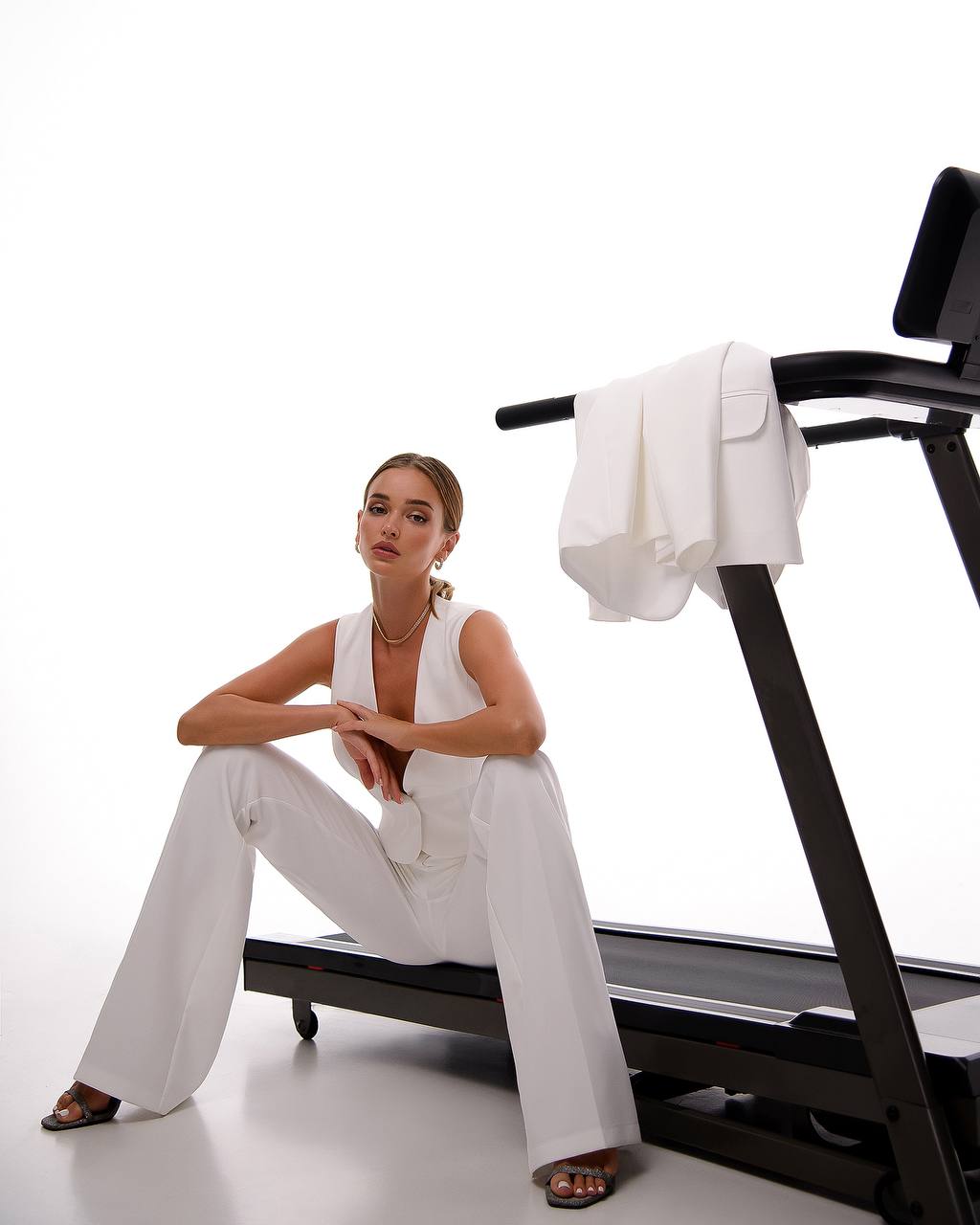 Woman in a white outfit sitting on a treadmill with a white towel draped over it against a white background