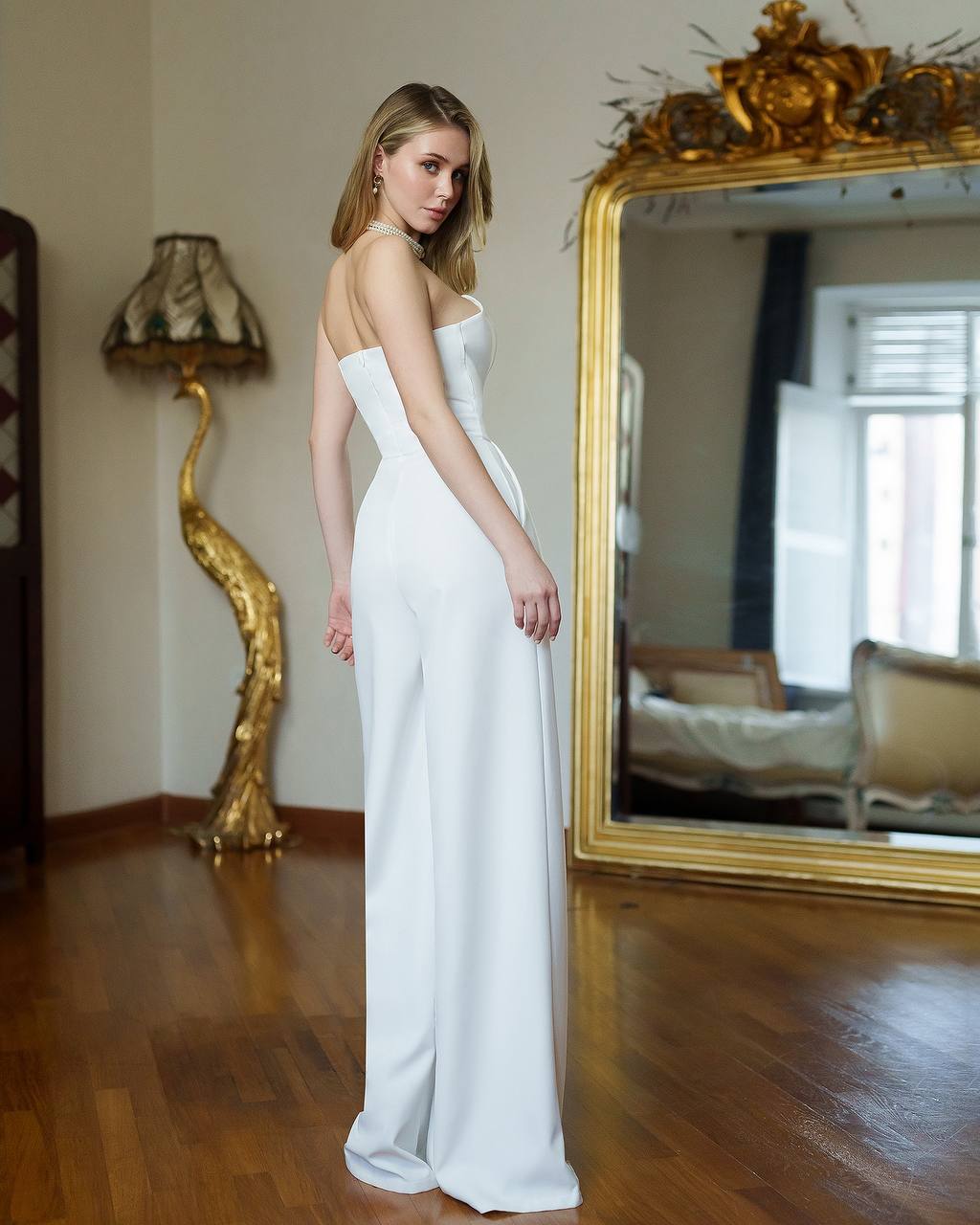 Woman in a white dress standing in a room with a large mirror and decorative lamp.
