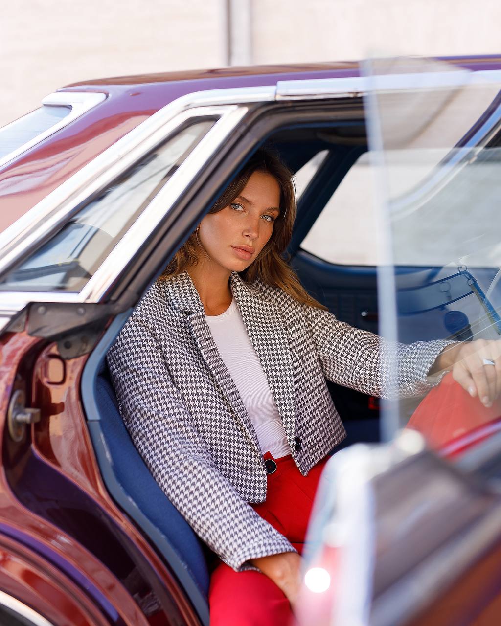 Woman sitting in a car wearing a checkered blazer and red pants.